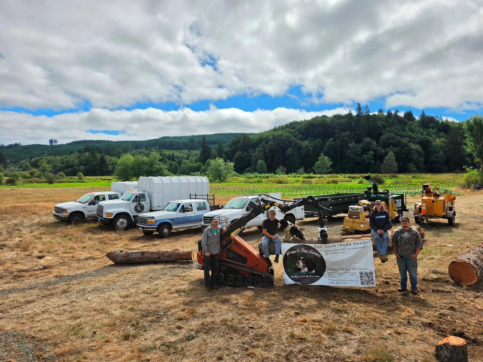 Four Womer’s crew members and a dog stand with work trucks, a compact loader, and tree-service equipment.